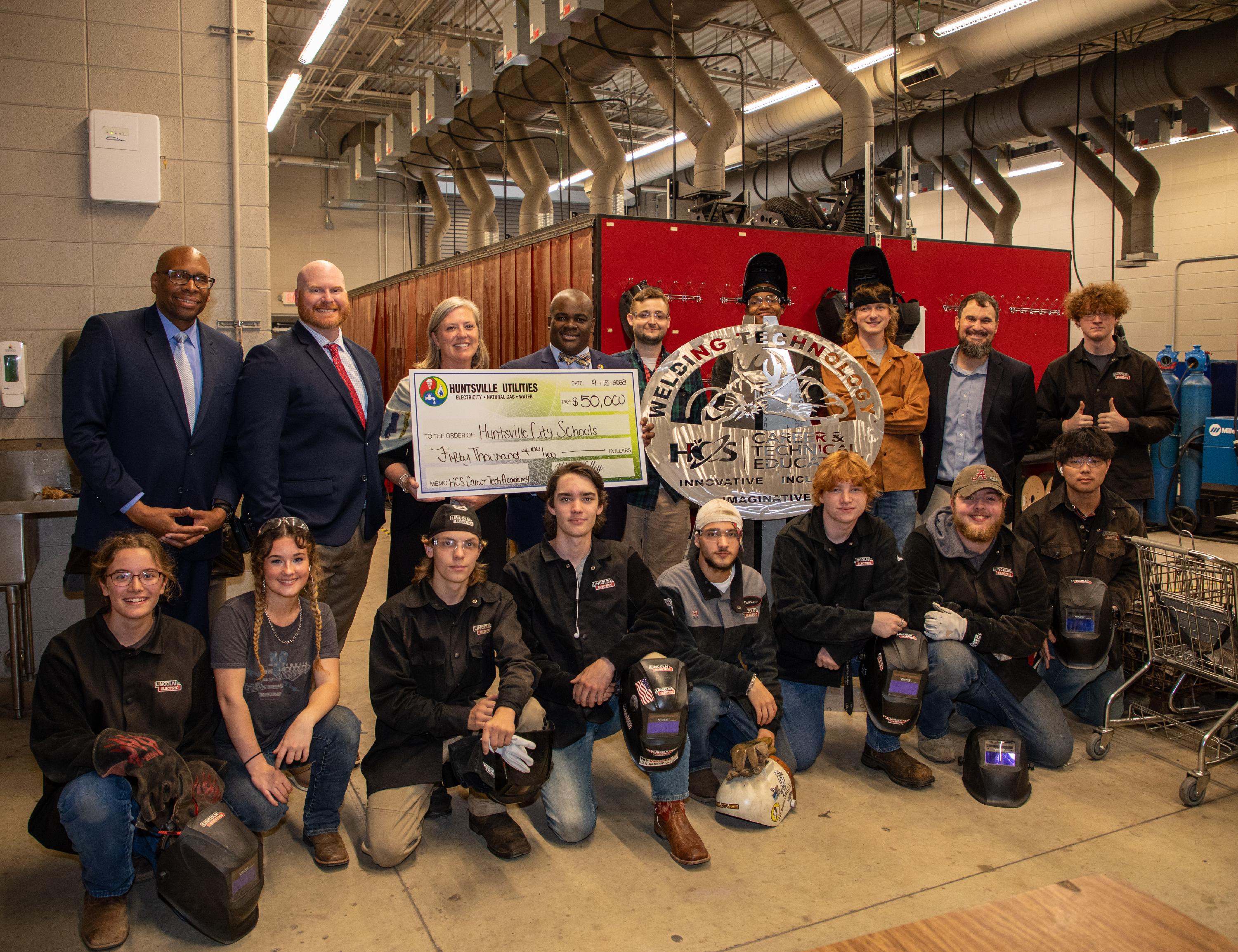 Group of welding students gathered around a metal sign they created. Huntsville City Schools and Huntsville Utilities officials holding large check for $50,000.